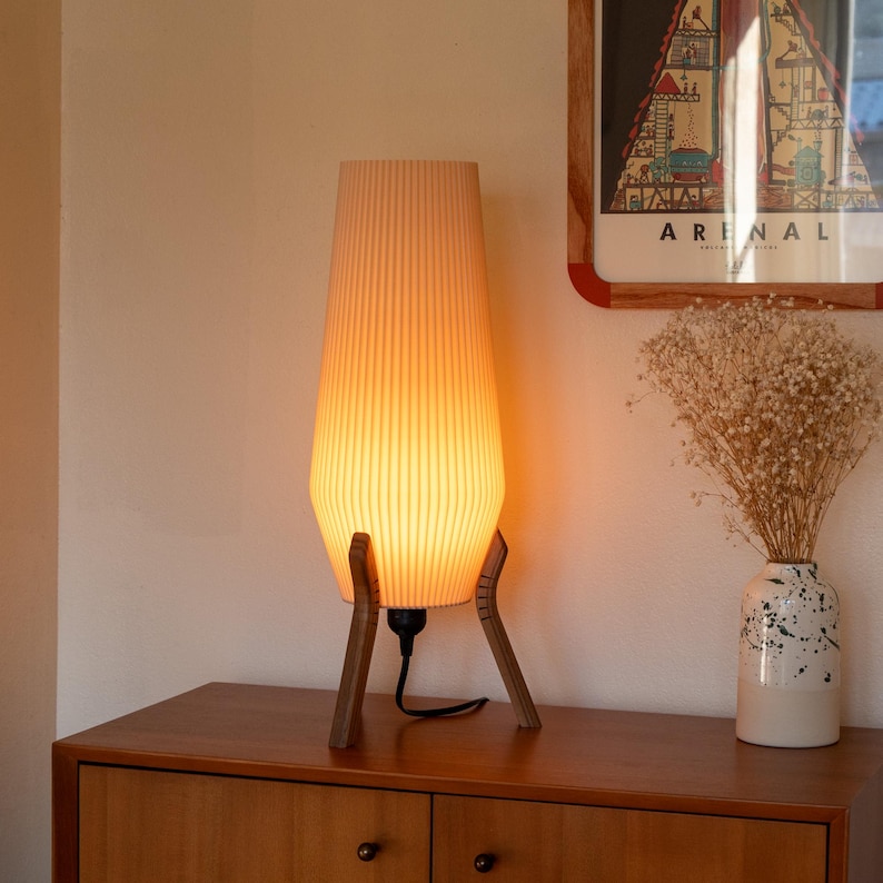 A mid-century modern table lamp with a cream-colored, vertically ribbed shade. The lamp is supported by a wooden tripod base. The lamp is lit, emitting a warm, inviting glow. The lamp is on a wooden cabinet.