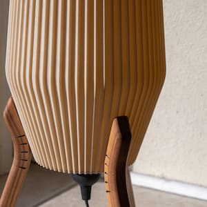 A beige, ribbed lampshade sits atop a wooden tripod base. The lamp's shade has a vertical, curved design. The wooden legs are dark brown and have a mid-century modern aesthetic. The lamp is a decorative lighting fixture.