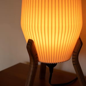A modern table lamp with a warm, glowing light. The lamp features a ribbed, off-white shade and is supported by a wooden tripod base. The shade emits a soft, inviting light, perfect for a living room or bedroom. The lamp's design is both stylish and functional.