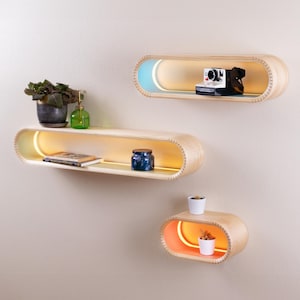 May include: Three oval-shaped wooden shelves with built-in LED lights. The top shelf is light blue, the middle shelf is white, and the bottom shelf is orange. The shelves are mounted on a white wall.