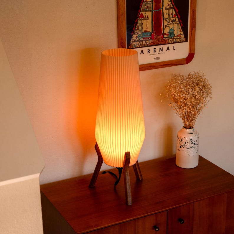 A mid-century modern style lamp with a warm, orange glow. The lamp has a ribbed, cylindrical shade and is supported by a tripod base made of dark wood. The lamp is placed on a dark wood cabinet, next to a vase of dried flowers.