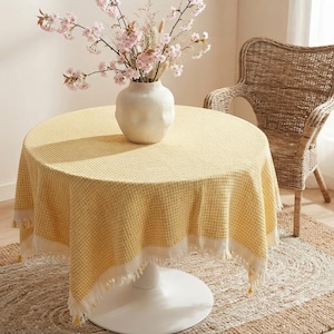 Peut inclure: Une table ronde recouverte d'une nappe jaune à texture gaufrée avec une bordure à franges blanches. Un vase en céramique blanche contenant des branches de cerisier en fleurs roses est posé au centre de la table, près d'une chaise en osier.