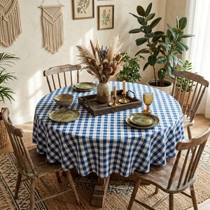 Peut inclure: Une table à manger ronde recouverte d'une nappe à carreaux bleus et blancs, avec un plateau en bois sombre contenant un vase en céramique rempli de fleurs séchées, deux bougeoirs en laiton, un bol en verre et deux assiettes disposées.