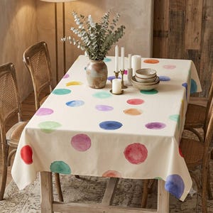 May include: A rectangular cream-coloured tablecloth featuring a scattered pattern of multicoloured watercolour-style polka dots in shades of red, blue, green, purple, and orange, draped over a wooden table with a vase and candles.