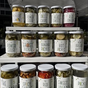 Peut inclure: Un présentoir d'étagère avec divers bocaux de légumes marinés et d'autres produits alimentaires. Les bocaux sont étiquetés avec des noms comme "The Power Sour", "The Ghost Pickle", "The Banana Pickle", "The Hot Pickle", "The New Bread Butter", "The Spicy Garlic", "The Red Jalapenos", "Sweet Jalapeno", et "The Pickle Juice".