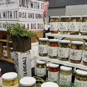Könnte beinhalten: Eine Auswahl an eingelegten Gemüsesorten in Gläsern mit weißen Etiketten. Die Gläser sind auf einem weißen Regal angeordnet, über dem ein Schild mit dem Text "DOLL WITH IT RISE AND BRINE" angebracht ist. Die Gläser sind mit verschiedenen Gurkenarten beschriftet, darunter "The Hot Pickle", "The New Bread Butter", "The Banana Pickle", "Spicy Spears", "Power Sour", "Heat Sweet", "Dill Chips", "The Ghost Pickle", "The Vintage Pickle", "Jalapeños", "Red Jalapeños", "The Banana Pepper" und "Sweet Pickle".