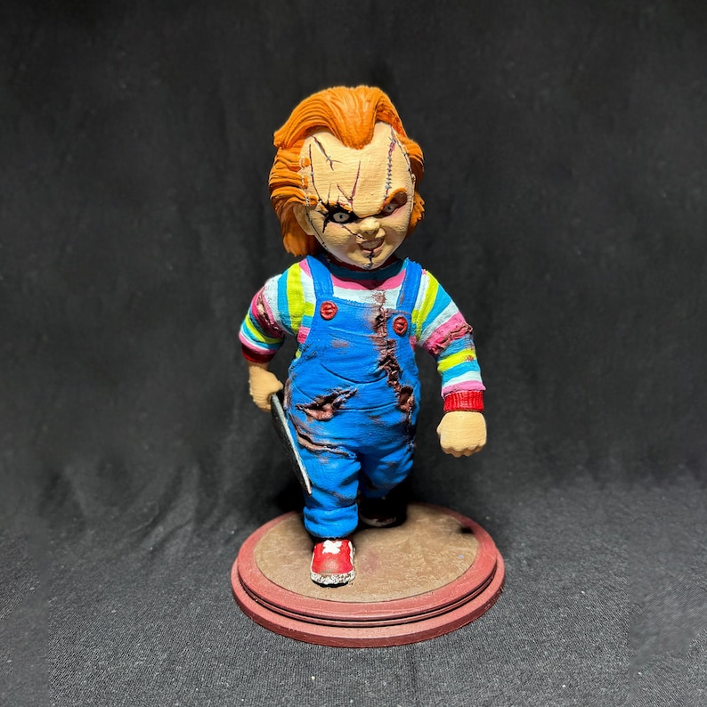 May include: A red-haired doll with a stitched face and a blue overall outfit holding a knife. The doll is standing on a brown circular base.