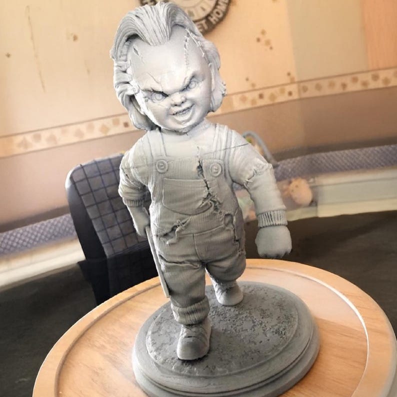 May include: A gray painted figurine of a fictional horror character, Chucky, from the Child's Play franchise. The figurine is standing on a round base and is holding a knife in his right hand.