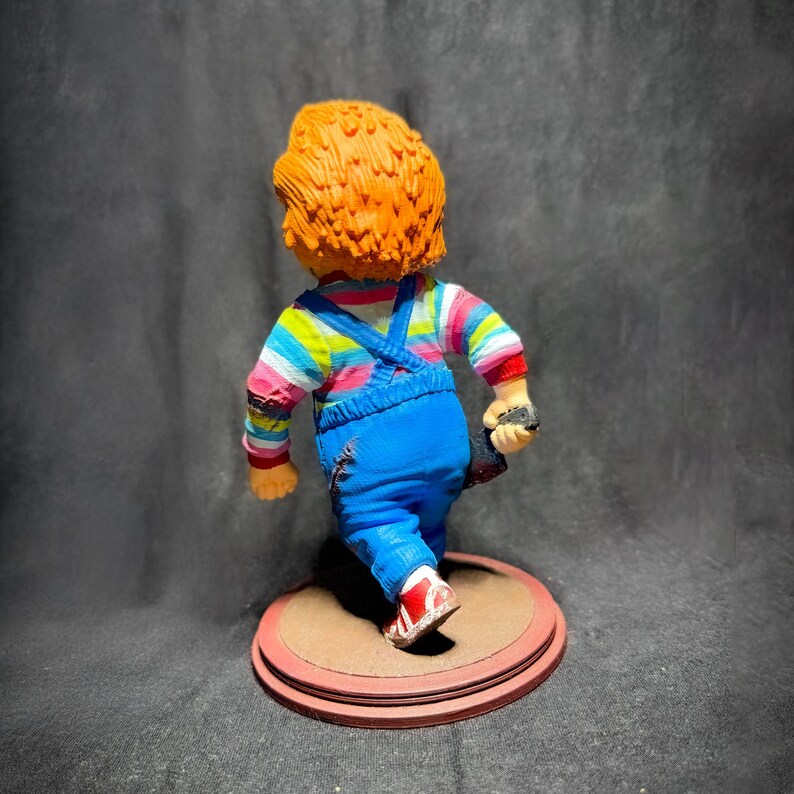 May include: A red-haired doll wearing a blue and white striped shirt, blue overalls, and red shoes. The doll is holding a knife and is standing on a brown base.