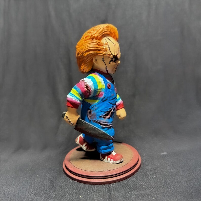 May include: A small, red-haired doll with a blue and white striped shirt and blue overalls. The doll is holding a black knife and has a menacing expression on its face. The doll is standing on a brown circular base.