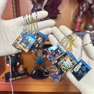 May include: Miniature PS4 game case keychains featuring game titles like Resident Evil 2, Grand Theft Auto, and The Last of Us Part II. Each keychain has a silver or gold-colored ring. The keychains are held up by gloved hands.