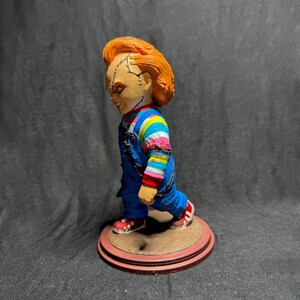 May include: A red-haired doll with a stitched face, wearing a blue and white striped shirt and blue overalls, stands on a brown and red base. The doll is a popular horror character.