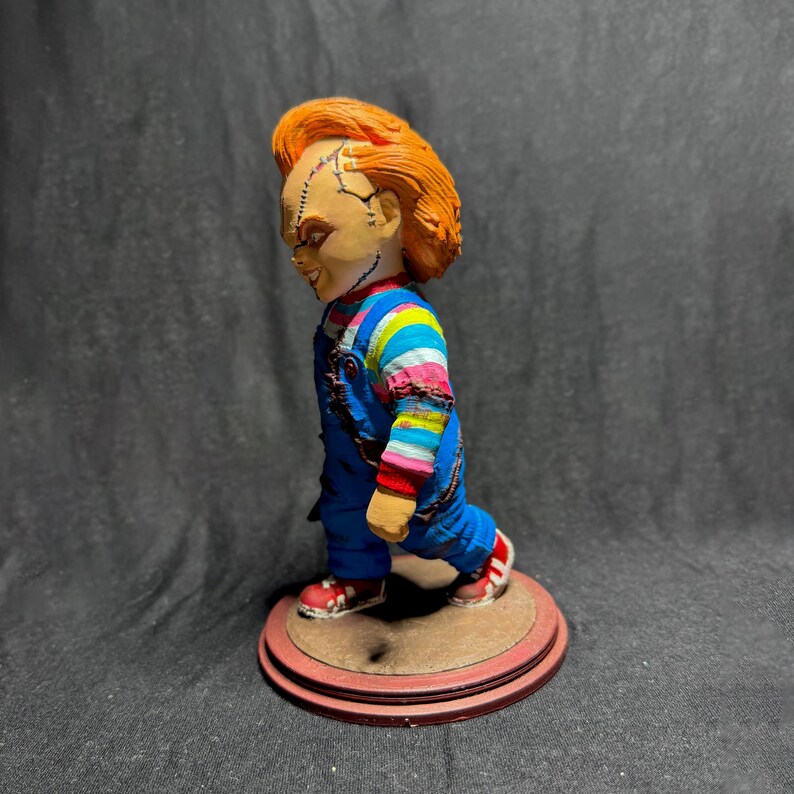 May include: A red-haired doll with a stitched face, wearing a blue and white striped shirt and blue overalls, stands on a brown and red base. The doll is a popular horror character.