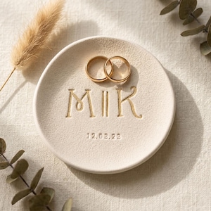 Pode incluir: Um prato redondo de cerâmica em tom off-white com as letras M e K em relevo separadas por duas linhas verticais, com a data 12.02.23 gravada abaixo. Dois anéis de metal dourado repousam sobre a superfície texturizada de acabamento fosco.