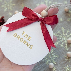 Puede incluir: Adorno redondo blanco con una cinta y un lazo rojos. El adorno tiene el texto "THE BROWNS" en letras doradas.