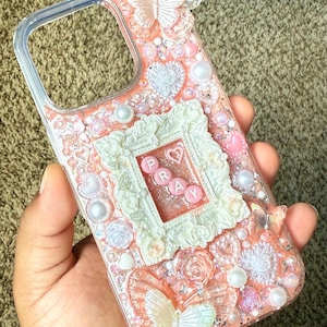 May include: A clear phone case with a pink and white decorative design. The case features butterflies, pearls, hearts, and the word "PRAY" spelled out in pink letters within a white frame.
