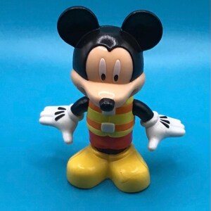 May include: A Mickey Mouse figurine with black ears, a black head, and a tan face. The figure wears a yellow and orange striped shirt, yellow shoes, and white gloves. The figure is standing with its arms outstretched.