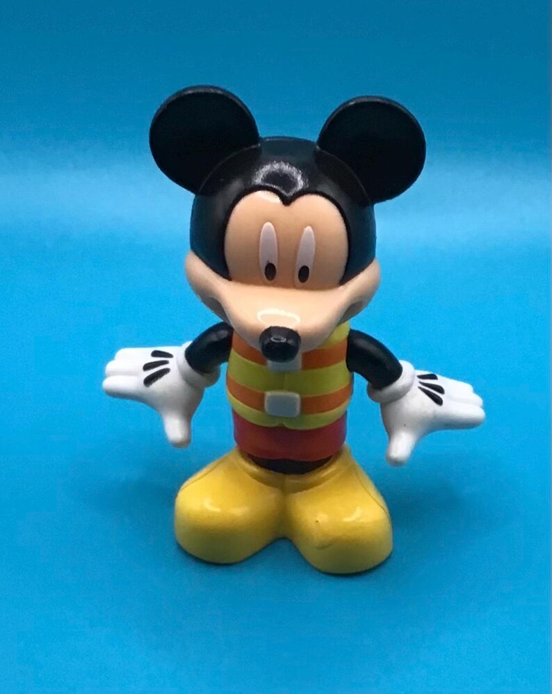 May include: A Mickey Mouse figurine with black ears, a black head, and a tan face. The figure wears a yellow and orange striped shirt, yellow shoes, and white gloves. The figure is standing with its arms outstretched.