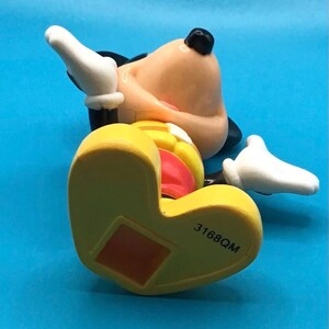 May include: A yellow, red, and black Mickey Mouse figurine lying on its back. The figurine has white gloves, a black nose, and a red tongue. The number "3168QM" is printed on the bottom of the yellow shoe.