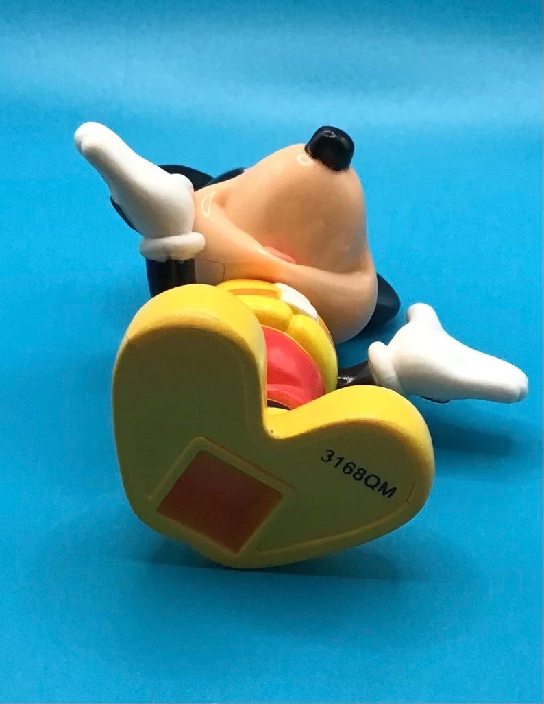 May include: A yellow, red, and black Mickey Mouse figurine lying on its back. The figurine has white gloves, a black nose, and a red tongue. The number "3168QM" is printed on the bottom of the yellow shoe.