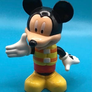 May include: A Mickey Mouse figurine with black ears, a tan face, and white gloves. He wears a yellow and orange striped vest over a red shirt and yellow shoes. The figure is posed with one hand up and the other down.
