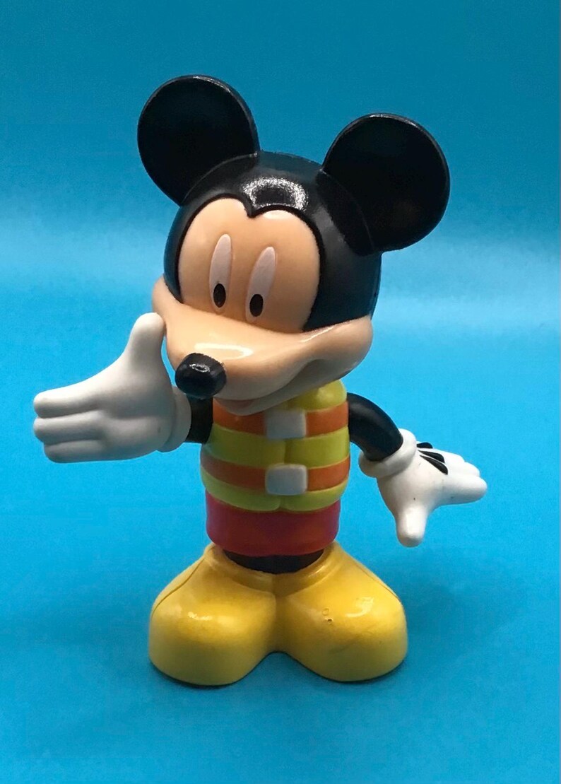 May include: A Mickey Mouse figurine with black ears, a tan face, and white gloves. He wears a yellow and orange striped vest over a red shirt and yellow shoes. The figure is posed with one hand up and the other down.