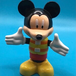 May include: A Mickey Mouse figurine with a black head and ears, a tan face, and white gloves. The figure wears a yellow and orange striped vest, red shorts, and yellow shoes. The background is a solid blue.