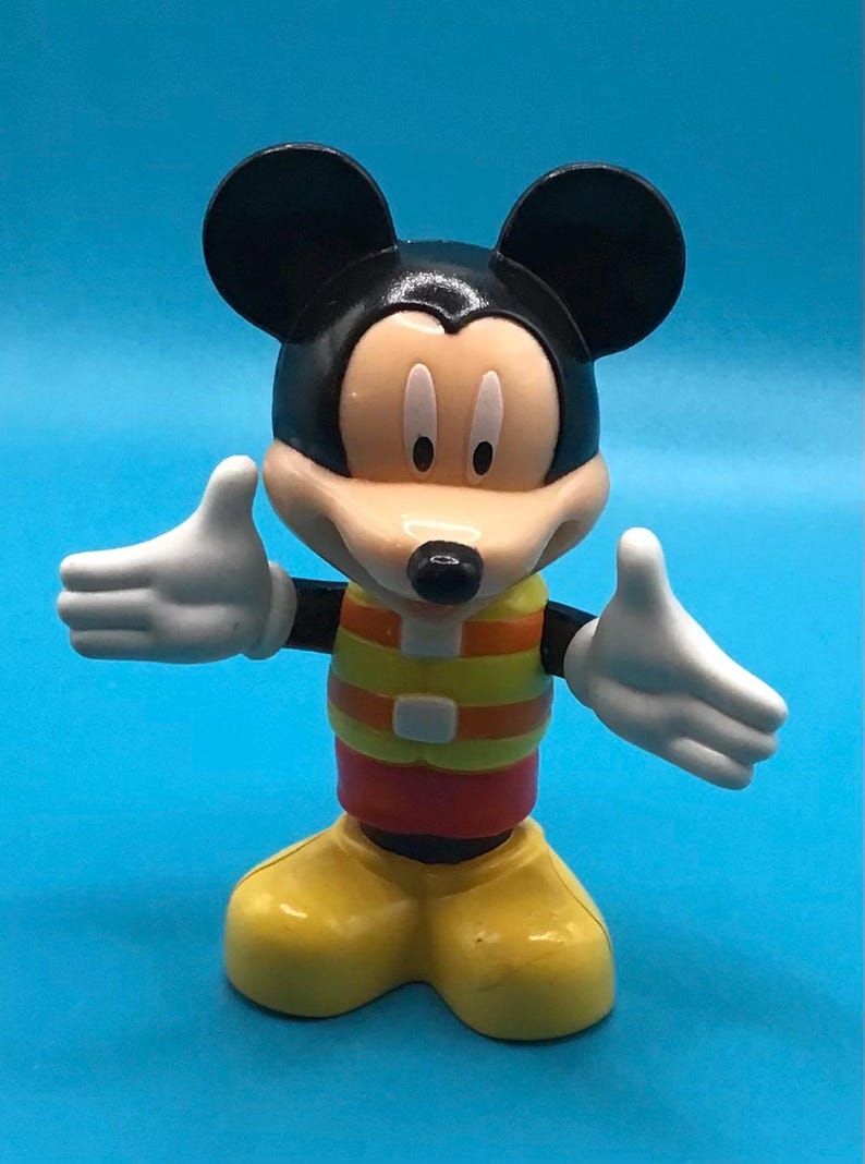 May include: A Mickey Mouse figurine with a black head and ears, a tan face, and white gloves. The figure wears a yellow and orange striped vest, red shorts, and yellow shoes. The background is a solid blue.