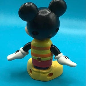 May include: A Mickey Mouse figurine, viewed from the back, with a black head and ears, a yellow and striped torso, and a yellow base. The figure has white gloves and is set against a blue background.