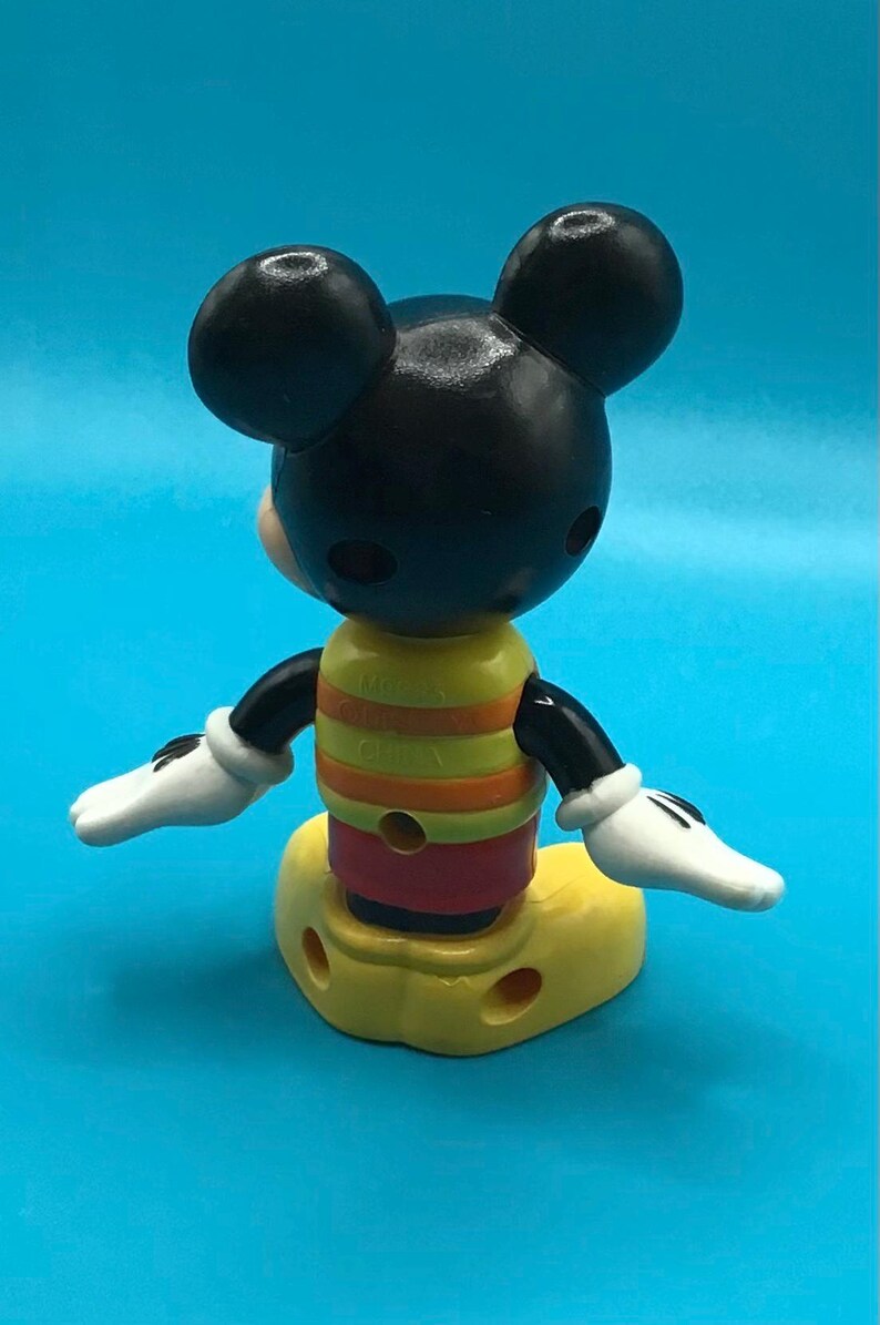 May include: A Mickey Mouse figurine, viewed from the back, with a black head and ears, a yellow and striped torso, and a yellow base. The figure has white gloves and is set against a blue background.