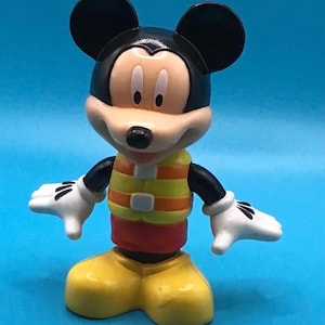 May include: A Mickey Mouse figurine, featuring black ears, a tan face, and a yellow and orange vest. The figure has white gloves, red shorts, and yellow shoes. The background is a solid blue.