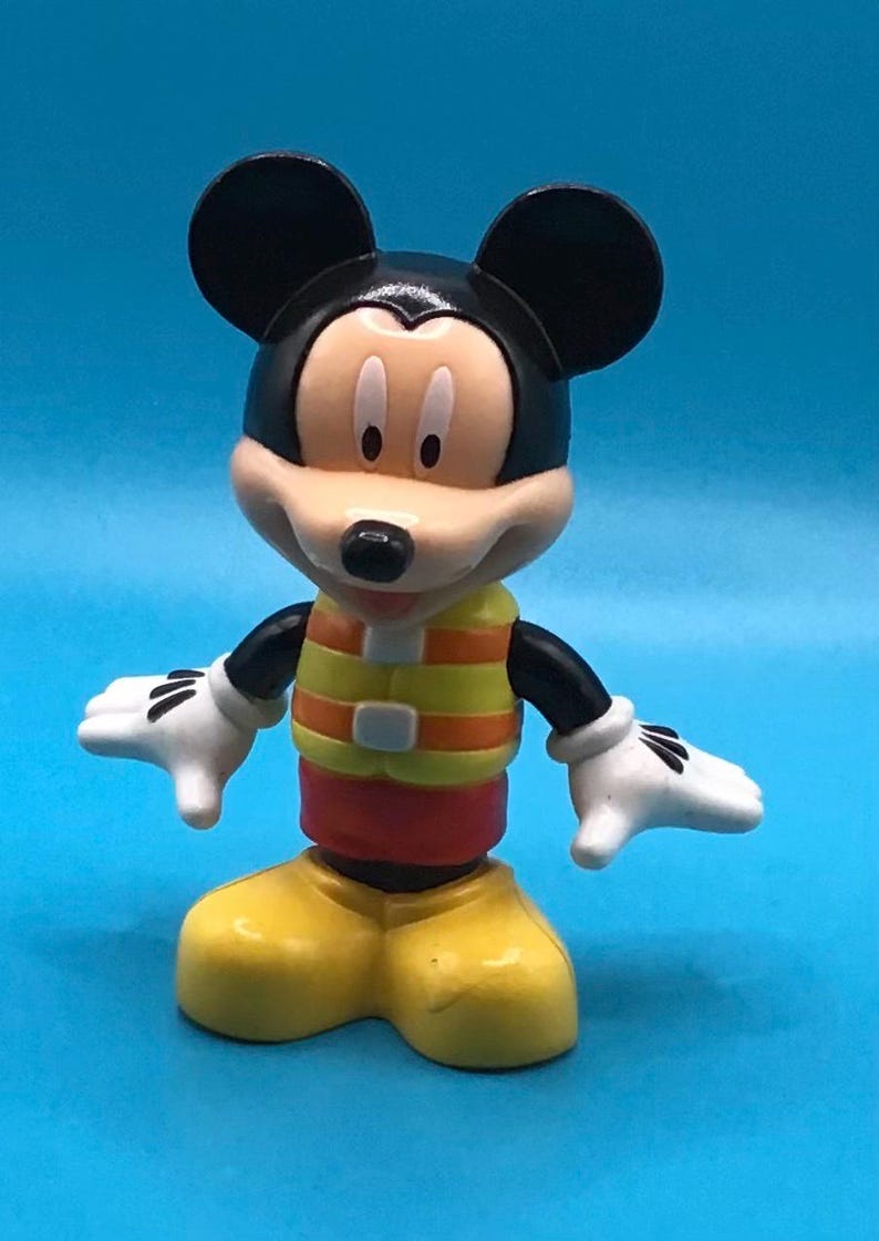 May include: A Mickey Mouse figurine, featuring black ears, a tan face, and a yellow and orange vest. The figure has white gloves, red shorts, and yellow shoes. The background is a solid blue.