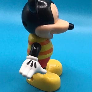 May include: A Mickey Mouse figurine, featuring a black head with large ears, a tan face, and a black nose. The figure wears a yellow and orange striped shirt, red shorts, white gloves, and yellow shoes. The background is a solid blue.