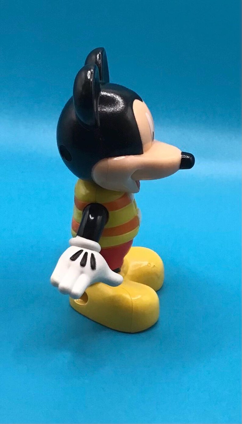 May include: A Mickey Mouse figurine, featuring a black head with large ears, a tan face, and a black nose. The figure wears a yellow and orange striped shirt, red shorts, white gloves, and yellow shoes. The background is a solid blue.
