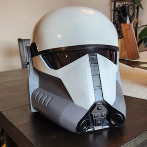 May include: A white and gray helmet with a black visor. The helmet has a textured surface and is sitting on a dark brown wooden table.