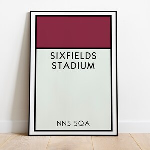 May include: A framed print of a minimalist design featuring a burgundy rectangle at the top, the text "SIXFIELDS STADIUM" in black, and the text "NN5 5QA" in black at the bottom.
