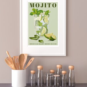 May include: A framed illustration of a mojito cocktail with the text "MOJITO" and "WHITE RUM | SUGAR | FRESH MINT | FRESH LIME | SODA WATER" on a green background. The illustration is surrounded by a white frame and is hanging on a light beige wall.