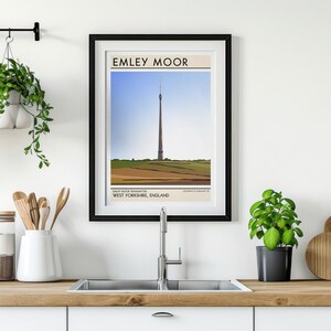 May include: A black framed poster with a white background and the text "Emley Moor" in black. The poster features a stylized illustration of a tall tower in a field with a blue sky in the background. The text "West Yorkshire, England" is below the illustration.