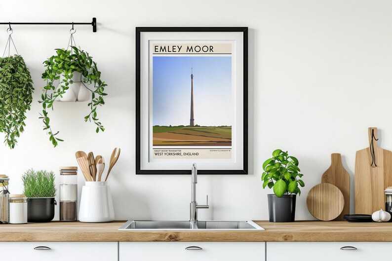 May include: A black framed poster with a white background and the text "Emley Moor" in black. The poster features a stylized illustration of a tall tower in a field with a blue sky in the background. The text "West Yorkshire, England" is below the illustration.