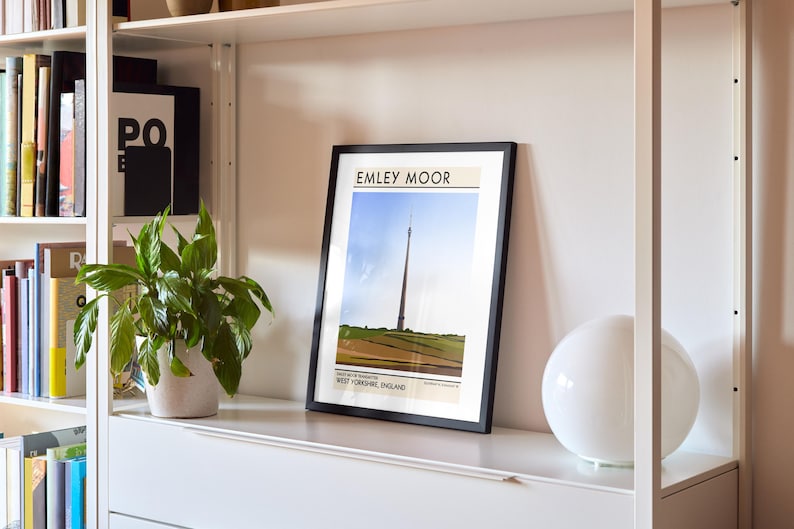 May include: A black framed print with the text "Emley Moor" and a picture of a tall tower in a field. The print is on a white shelf with a white lamp and a potted plant.