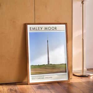 May include: A framed print of a minimalist illustration of the Emley Moor Transmitter, a tall tower in West Yorkshire, England. The print has a white background with black text and a brown frame.