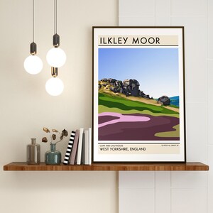 May include: A minimalist poster with a brown frame featuring a colorful illustration of Cow and Calf Rocks in Ilkley Moor, West Yorkshire, England. The poster has the text "ILKLEY MOOR" in a bold, sans-serif font at the top and "COW AND CALF ROCKS WEST YORKSHIRE, ENGLAND" at the bottom.
