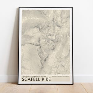 May include: A framed topographic map print of Scafell Pike in the Lake District, Southern Fells, England. The print features beige and grey contour lines, with the text "LAKE DISTRICT, SOUTHERN FELLS, ENGLAND" and "SCAFELL PIKE" at the bottom.