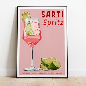 May include: A framed poster featuring a pink background with a Sarti Spritz cocktail in a stemmed glass, garnished with a lime slice and ice cubes. The text reads SARTI Spritz and lists ingredients: SARTI ROSA, PROSECCO, SODA WATER, LIME SLICE, CUBED ICE.