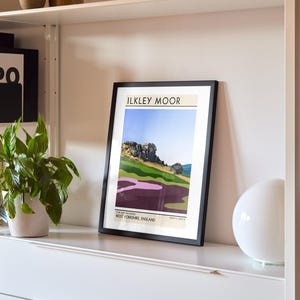 May include: A framed print of a landscape illustration with the text "ILKLEY MOOR" and "WEST YORKSHIRE, ENGLAND" on a white shelf with a white lamp.