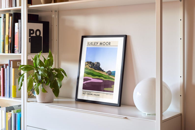 May include: A framed print of a landscape illustration with the text "ILKLEY MOOR" and "WEST YORKSHIRE, ENGLAND" on a white shelf with a white lamp.