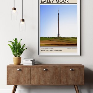May include: A minimalist poster with a brown background and white text that reads "EMLEY MOOR". The poster features a stylized illustration of a tall, slender tower against a blue sky and green hills. The text "EMLEY MOOR TRANSMITTER WEST YORKSHIRE, ENGLAND" is printed below the illustration.