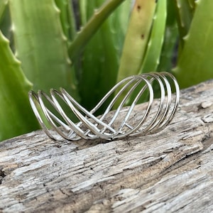 Peut inclure: Un bracelet en argent composé de plusieurs bandes fines entrelacées. Le bracelet est posé sur un morceau de bois usé.