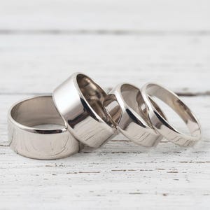 Può includere: Quattro anelli d'argento di diverse larghezze sono disposti su una superficie di legno bianco. Gli anelli hanno una superficie lucida e riflettente. L'anello più largo è a sinistra, il più sottile a destra. Gli anelli sono probabilmente in metallo.