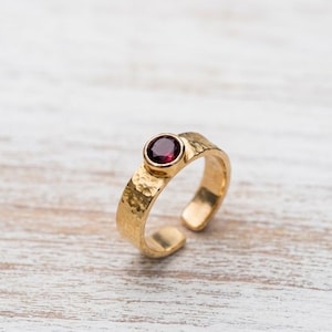 Könnte beinhalten: Ein goldfarbener Ring mit einem gehämmerten, strukturierten Band und einem offenen Design auf der Rückseite. Der Ring ist mit einem einzelnen, runden, tiefroten Edelstein in einer glatten, kreisförmigen Zargenfassung besetzt.